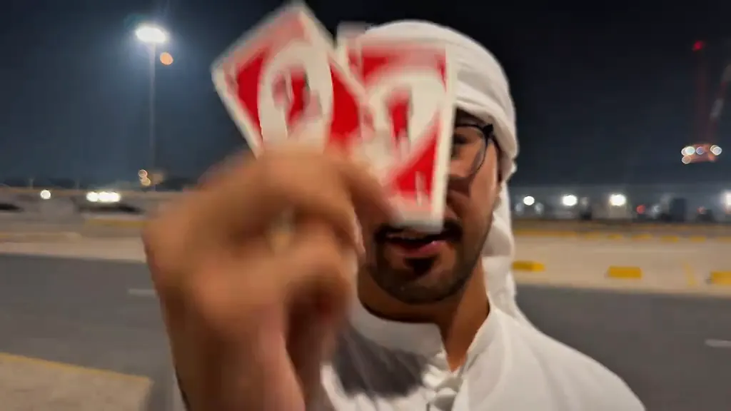 A man sweeps two red cards toward the lens in a floodlit parking lot at night
